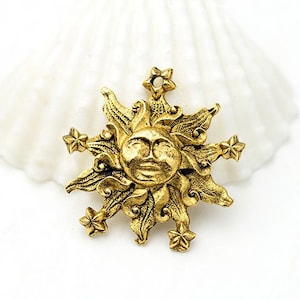 Puede incluir: Colgante de sol dorado con una cara sonriente y detalles de estrellas. El sol tiene rayos detallados y está rodeado de seis pequeñas estrellas. El colgante es probablemente de metal y tiene una estética antigua o vintage.