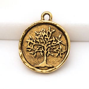 May include: A round, antique gold-colored charm featuring a detailed tree design. The tree has a prominent trunk and branches with leaves, set within a circular frame. The charm has a loop at the top for attaching to jewelry.