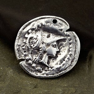 May include: A silver-colored coin pendant with a detailed relief of a person in profile wearing a helmet. The coin has a distressed, antique appearance with a hole at the top for stringing.