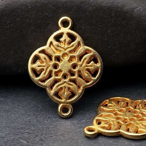 May include: Two gold-coloured filigree connectors with a four-leaf clover design. Each connector has a central circular design with intricate cut-out patterns and small loops for attachment. The connectors are displayed on a dark grey surface.