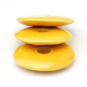 Puede incluir: Tres objetos amarillos en forma de rosquilla apilados, con un agujero en el centro y un acabado brillante. Se colocan sobre un fondo blanco, creando una composición minimalista y limpia.