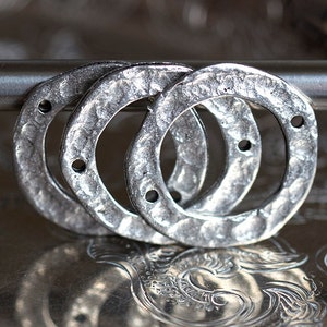 Könnte beinhalten: Nahaufnahme von drei silberfarbenen, gehämmerten Metallringen mit kleinen Löchern. Die Ringe sind gestapelt und leicht überlappend, mit einer strukturierten Oberfläche. Die Ringe sind wahrscheinlich für die Schmuckherstellung oder Bastelprojekte gedacht.