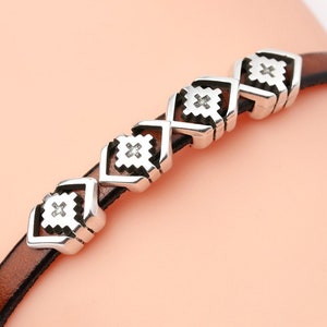 May include: A brown leather bracelet with silver-colored decorative beads. The beads are square-shaped with an X design in the center. The bracelet is shown on a person's wrist.