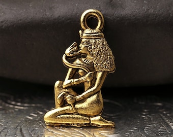 May include: A gold-colored pendant featuring an Egyptian figure with intricate details. The figure is kneeling and holding a snake. The pendant has a loop at the top for attaching to a chain or cord. The overall design is reminiscent of ancient Egyptian art.