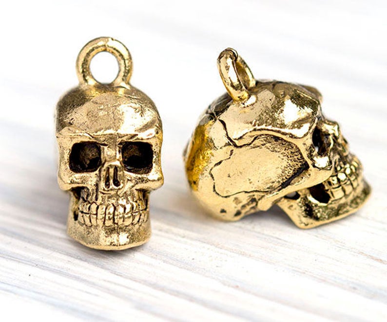 May include: Two gold-colored skull charms. One is upright, the other is on its side. Both have a loop at the top for hanging. The skulls have detailed features, including eye sockets, a nose cavity, and teeth. The charms have a metallic finish.