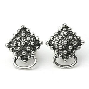 May include: Pair of silver-toned stud earrings. Each earring features a diamond-shaped design covered in small, raised dots. A small, curved loop hangs below the diamond shape. The earrings are set against a white background.