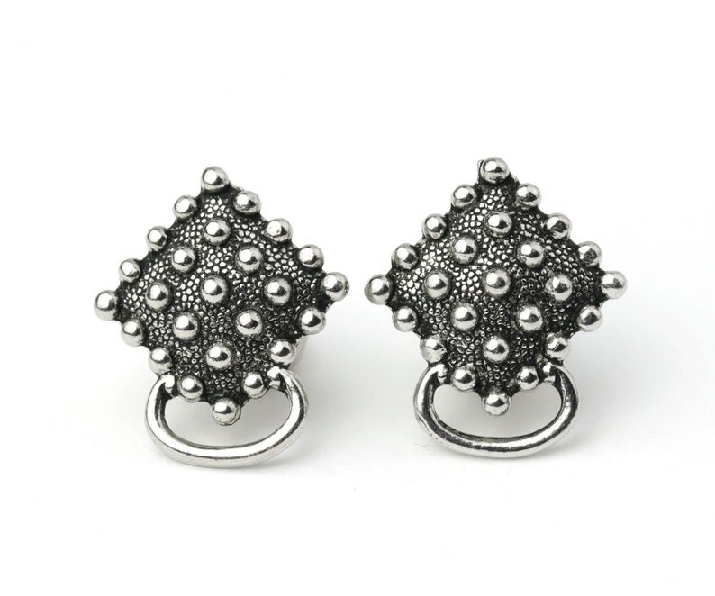 May include: Pair of silver-toned stud earrings. Each earring features a diamond-shaped design covered in small, raised dots. A small, curved loop hangs below the diamond shape. The earrings are set against a white background.