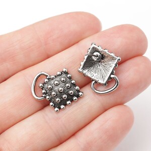 May include: A pair of silver-toned stud earrings. Each earring features a square design with a textured surface of small, raised dots. A small loop extends from the bottom of each earring. The earrings are displayed against a light background.