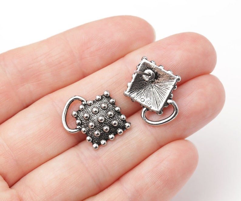 May include: A pair of silver-toned stud earrings. Each earring features a square design with a textured surface of small, raised dots. A small loop extends from the bottom of each earring. The earrings are displayed against a light background.