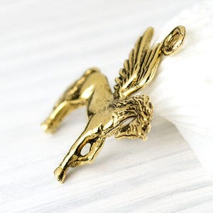 Op de afbeelding: Een gedetailleerde, antiek goudkleurige Pegasus bedel. Het mythische gevleugelde paard is afgebeeld in een dynamische pose, met uitgestrekte vleugels en een vloeiende manen. De bedel heeft een lus aan de bovenkant voor bevestiging.