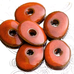 May include: Six reddish-brown, oval-shaped beads with a central hole. The beads have a glossy finish and a slightly textured surface. They are arranged on a silver, patterned surface. These beads could be used for jewelry making or other craft projects.