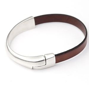 Peut inclure: Un bracelet en cuir marron avec un fermoir argenté. Le bracelet présente un design minimaliste, avec un élément métallique incurvé relié au bracelet en cuir. Le bracelet en cuir mesure environ 0,5 cm de large.