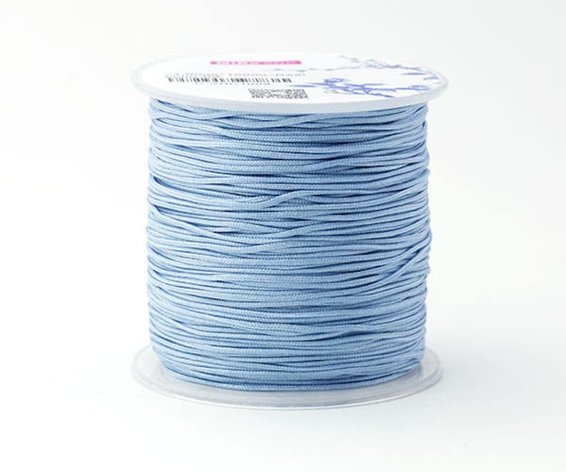 Puede incluir: Un carrete de cord&oacute;n azul claro, enrollado firmemente y exhibido sobre un fondo blanco. El cord&oacute;n es de color azul claro liso, y el carrete tiene una base de pl&aacute;stico transparente. La parte superior del carrete tiene una etiqueta blanca con texto y dise&ntilde;os florales.