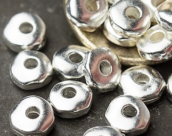 May include: Close-up of several shiny, silver-colored metal beads. The beads are irregularly shaped with a hole in the center, and are scattered on a dark surface. Some beads are in a small, silver spoon.