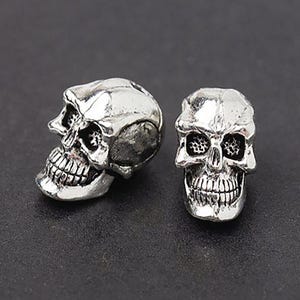 Puede incluir: Dos cuentas de calavera de color plateado. Una cuenta est&aacute; de perfil, mostrando el lado de la calavera, la otra est&aacute; orientada hacia adelante. Las calaveras tienen detalles, incluyendo cuencas de los ojos, cavidad nasal y dientes. Estas cuentas se utilizan para hacer joyas.