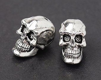 May include: Two silver-colored skull beads. One bead is positioned in profile, showcasing the side of the skull, while the other faces forward. The skulls have detailed features, including eye sockets, a nose cavity, and teeth. These beads are likely used for jewelry making.