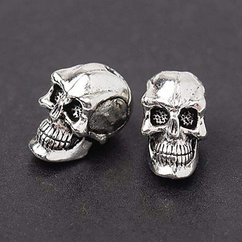 Puede incluir: Dos cuentas de calavera de color plateado. Una cuenta est&aacute; de perfil, mostrando el lado de la calavera, la otra est&aacute; orientada hacia adelante. Las calaveras tienen detalles, incluyendo cuencas de los ojos, cavidad nasal y dientes. Estas cuentas se utilizan para hacer joyas.