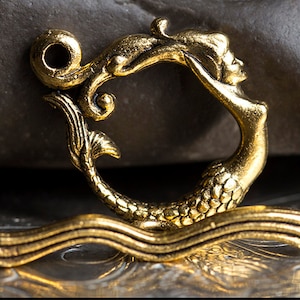 May include: An antique brass mermaid charm. The charm features a mermaid with flowing hair and a fishtail, set against a wave-like base. It has a loop at the top for hanging and is suitable for jewellery making or crafting projects.