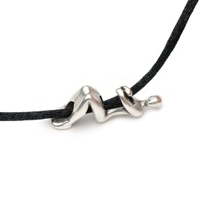 May include: A silver-colored pendant in the shape of a reclining figure strung on a black cord. The abstract design features smooth curves and a minimalist aesthetic, suitable for everyday wear.