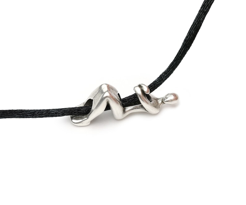 May include: A silver-colored pendant in the shape of a reclining figure strung on a black cord. The abstract design features smooth curves and a minimalist aesthetic, suitable for everyday wear.