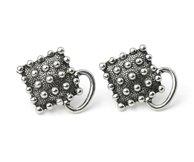 May include: A pair of silver-toned stud earrings. Each earring features a textured, square-shaped design covered in small, raised spheres. A curved, handle-like element extends from the side of each earring, creating a unique, artistic look.