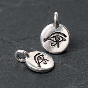 Peut inclure: Deux pendentifs argent&eacute;s repr&eacute;sentant le symbole de l'&OElig;il d'Horus. L'un est rond, l'autre ovale. L'&OElig;il d'Horus est un &oelig;il noir stylis&eacute; avec des d&eacute;tails complexes. Les deux pendentifs ont une petite boucle en haut pour attacher une cha&icirc;ne ou un cordon.