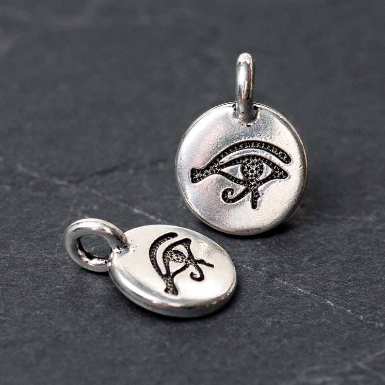 Peut inclure: Deux pendentifs argent&eacute;s repr&eacute;sentant le symbole de l'&OElig;il d'Horus. L'un est rond, l'autre ovale. L'&OElig;il d'Horus est un &oelig;il noir stylis&eacute; avec des d&eacute;tails complexes. Les deux pendentifs ont une petite boucle en haut pour attacher une cha&icirc;ne ou un cordon.