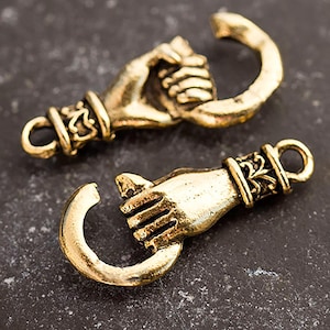 May include: Two antique gold-coloured clasps, each featuring a hand grasping a curved hook. The clasps have intricate detailing around the wrist and the hook's base. The background is a dark, textured surface.