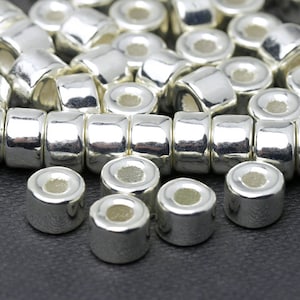 May include: Close-up of shiny silver metal beads. The beads are cylindrical with a slightly rounded shape and a hole running through the center. The beads are arranged in rows, suggesting they are for jewelry making or crafts.