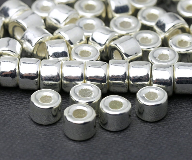 May include: Close-up of shiny silver metal beads. The beads are cylindrical with a slightly rounded shape and a hole running through the center. The beads are arranged in rows, suggesting they are for jewelry making or crafts.