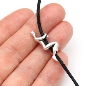 May include: A silver-colored pendant in the shape of a person climbing a rope. The pendant is attached to a black cord. The image is set against a white background.
