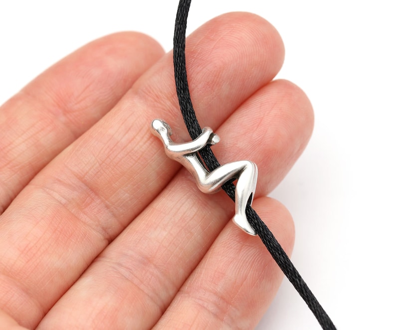 May include: A silver-colored pendant in the shape of a person climbing a rope. The pendant is attached to a black cord. The image is set against a white background.