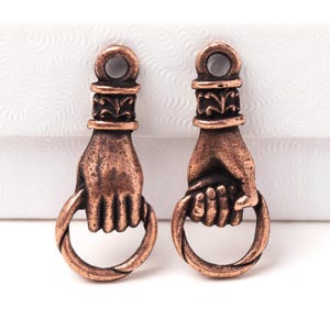 Puede incluir: Dos colgantes en forma de mano de color bronce, cada uno sosteniendo un anillo circular. Los colgantes tienen un diseño detallado en la mano y una parte superior decorativa con un lazo para colgar. El fondo es blanco.
