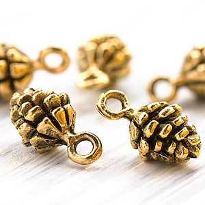 May include: Close-up of several small, gold-coloured pinecone charms. Each charm has a detailed, textured surface and a loop for attaching to jewellery or other crafts. The charms are arranged on a white, wooden surface.