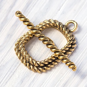 May include: A gold-colored toggle clasp with a rope design. The clasp features a circular loop and a bar that twists through the loop to secure it. The metal has a textured, aged appearance, suggesting a vintage or nautical theme.