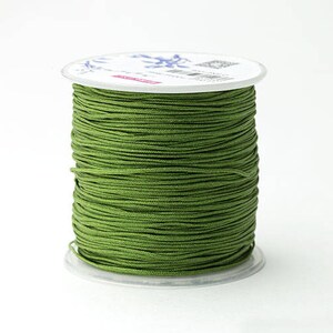Puede incluir: Una bobina de cord&oacute;n verde oliva, bien enrollada, se asienta sobre una base de pl&aacute;stico transparente. El cord&oacute;n es de color liso y la bobina tiene una parte superior blanca con detalles impresos. El fondo es blanco liso.