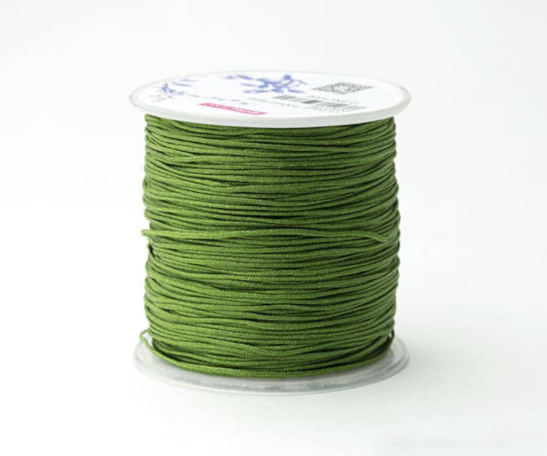 Puede incluir: Una bobina de cord&oacute;n verde oliva, bien enrollada, se asienta sobre una base de pl&aacute;stico transparente. El cord&oacute;n es de color liso y la bobina tiene una parte superior blanca con detalles impresos. El fondo es blanco liso.