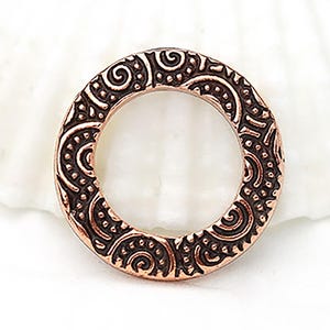 Puede incluir: Un colgante circular de color cobre con un diseño en relieve intrincado. El patrón presenta líneas onduladas, puntos y formas curvas, creando una apariencia ornamental detallada. El colgante tiene una abertura central y un borde ligeramente elevado.