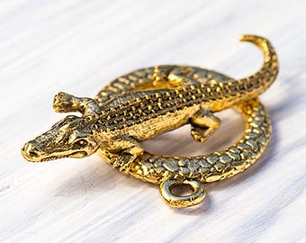 May include: A gold-colored alligator clasp, featuring a detailed reptile design. The alligator is positioned over a circular ring, with a textured surface. The clasp is likely used for jewelry or accessories.