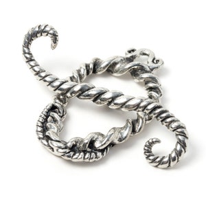 May include: A silver-colored, stylized metal object with a twisted rope design. The object has a looping, cursive shape with curved ends, and appears to be a decorative element or jewelry piece. The metal has a slightly aged or antique appearance.