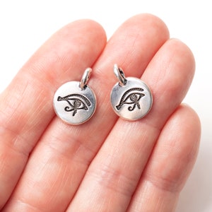 Peut inclure: Deux petits pendentifs ronds de couleur argent&eacute;e. Chaque pendentif pr&eacute;sente un symbole grav&eacute; de l'&OElig;il d'Horus. Les pendentifs ont une petite boucle en haut pour les attacher &agrave; une cha&icirc;ne ou &agrave; d'autres bijoux. Les pendentifs sont tenus dans une main.