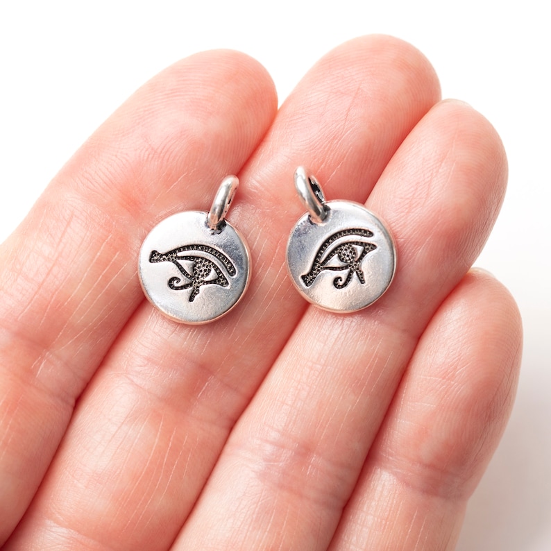 Peut inclure: Deux petits pendentifs ronds de couleur argent&eacute;e. Chaque pendentif pr&eacute;sente un symbole grav&eacute; de l'&OElig;il d'Horus. Les pendentifs ont une petite boucle en haut pour les attacher &agrave; une cha&icirc;ne ou &agrave; d'autres bijoux. Les pendentifs sont tenus dans une main.