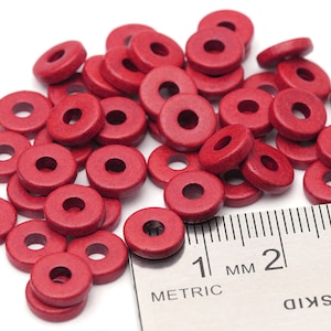 May include: A pile of small, red, wooden, ring-shaped beads. A metric ruler is next to the beads for scale. The beads are scattered on a white surface.