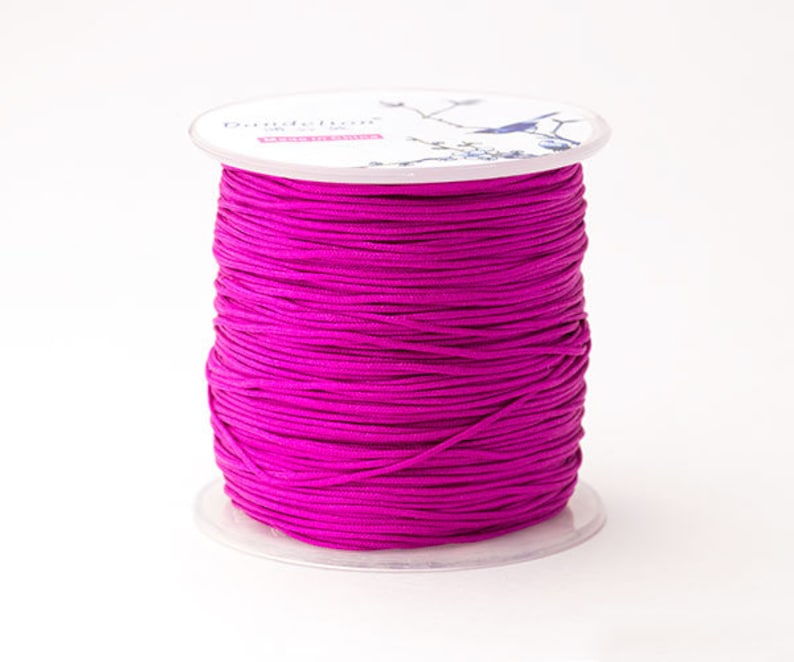Puede incluir: Un carrete de cord&oacute;n magenta vibrante. El cord&oacute;n est&aacute; enrollado alrededor de una bobina de pl&aacute;stico transparente. La parte superior de la bobina tiene una etiqueta blanca con el nombre de la marca "Dandelion" y un dise&ntilde;o decorativo de p&aacute;jaro. El cord&oacute;n parece liso y es adecuado para manualidades.