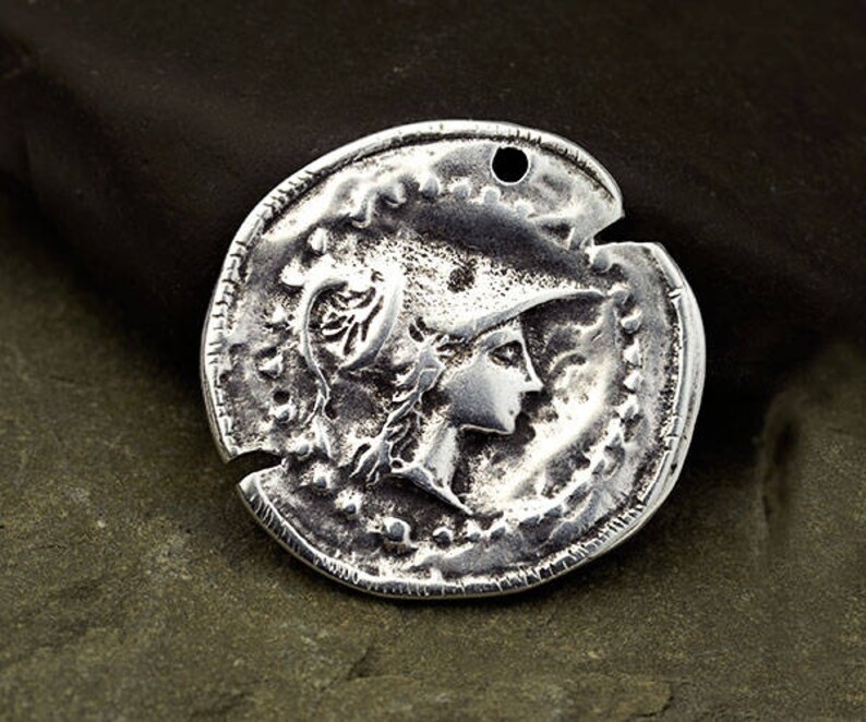 May include: A silver-colored coin pendant with a detailed relief of a person in profile wearing a helmet. The coin has a distressed, antique appearance with a hole at the top for stringing.