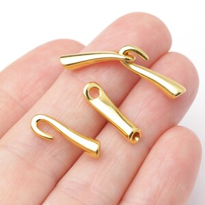 May include: Four small, shiny gold-colored metal clasps. Each clasp has a curved hook on one end and a small hole or opening on the other. The clasps are smooth and have a polished finish, reflecting light. They appear to be jewelry findings or components.