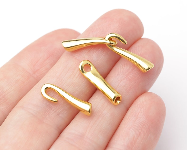May include: Four small, shiny gold-colored metal clasps. Each clasp has a curved hook on one end and a small hole or opening on the other. The clasps are smooth and have a polished finish, reflecting light. They appear to be jewelry findings or components.