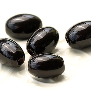 May include: A collection of glossy, black olives. The olives are oval in shape and have a smooth, reflective surface. They are presented on a white background, with some showing a small hole at one end. The olives look fresh and ready to eat.