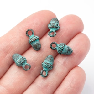 May include: Five small acorn charms with a distressed, aged appearance. The charms are a mix of teal and brown, with a loop at the top for attaching to jewellery or other crafts. The acorn caps have a textured pattern.