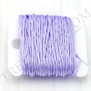 Puede incluir: Un carrete de cord&oacute;n de sat&eacute;n de color lavanda. El cord&oacute;n est&aacute; enrollado firmemente en un carrete de pl&aacute;stico blanco. La textura del cord&oacute;n es suave y parece estar hecho de muchos hilos finos. El carrete est&aacute; sobre un fondo blanco.